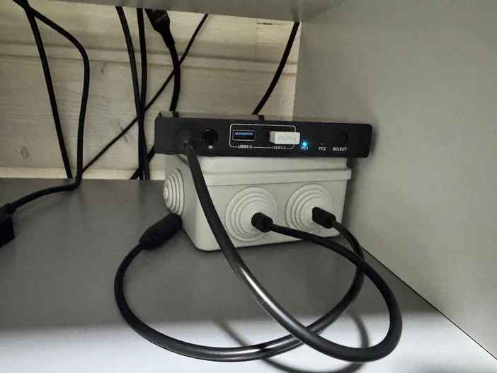 Un boîtier blanc avec des câbles noirs connectés à un commutateur USB sous une étagère.